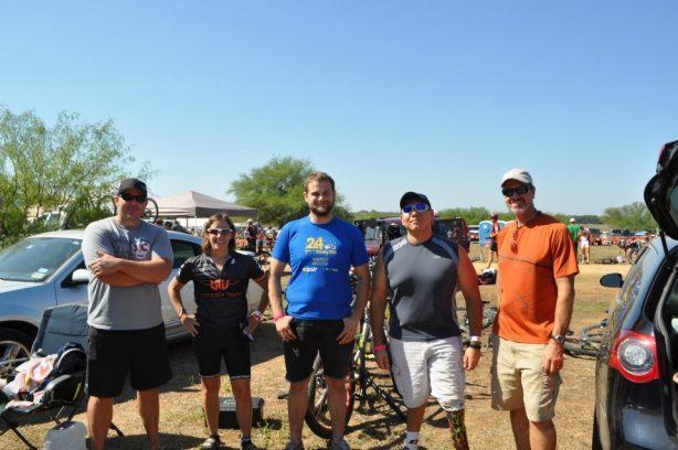 A group of five people standing together outdoors at an event, with a clear blue sky in the background. They are smiling and dressed in casual athletic attire, with bicycles and tents visible in the background, suggesting a sporting or recreational gathering. Rocky Hill Ranch mountain bike trail.