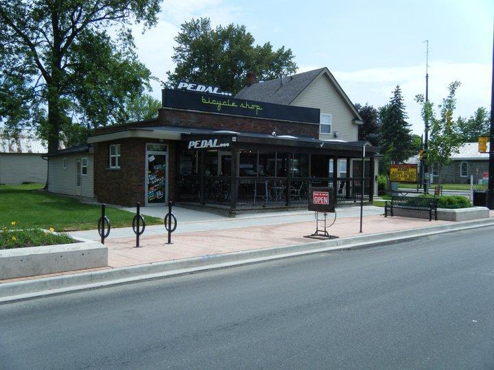 A bicycle shop named "PEDAL" with a large sign displaying the name on the roof. The shop features a patio area with bicycles on display inside and outside. There is a sign indicating that the shop is open, along with bike racks in front and benches nearby. The surrounding area includes trees and a well-maintained sidewalk.