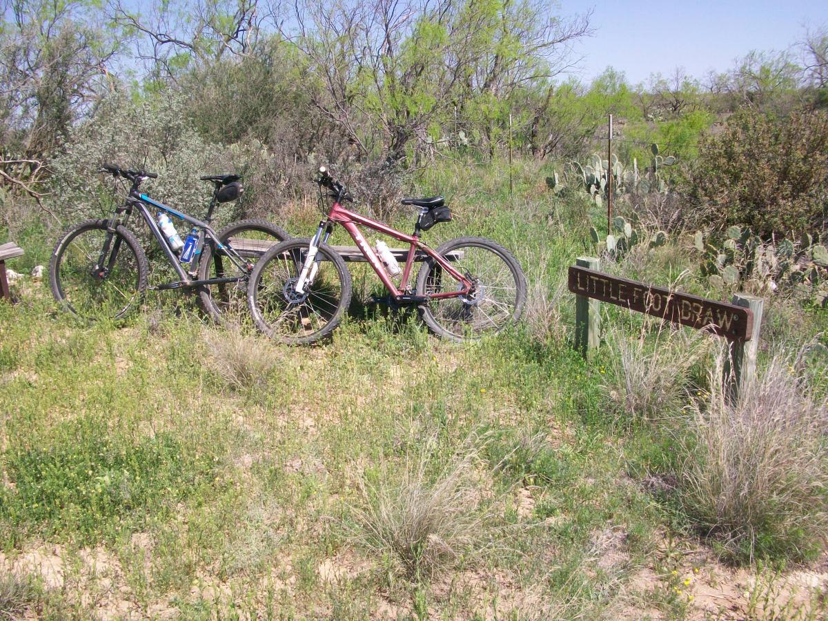 Two mountain bikes are parked beside a sign that reads "Little Foot Draw," surrounded by green grass, shrubs, and cacti in a rural landscape. San Angelo Trail mountain bike trail.
