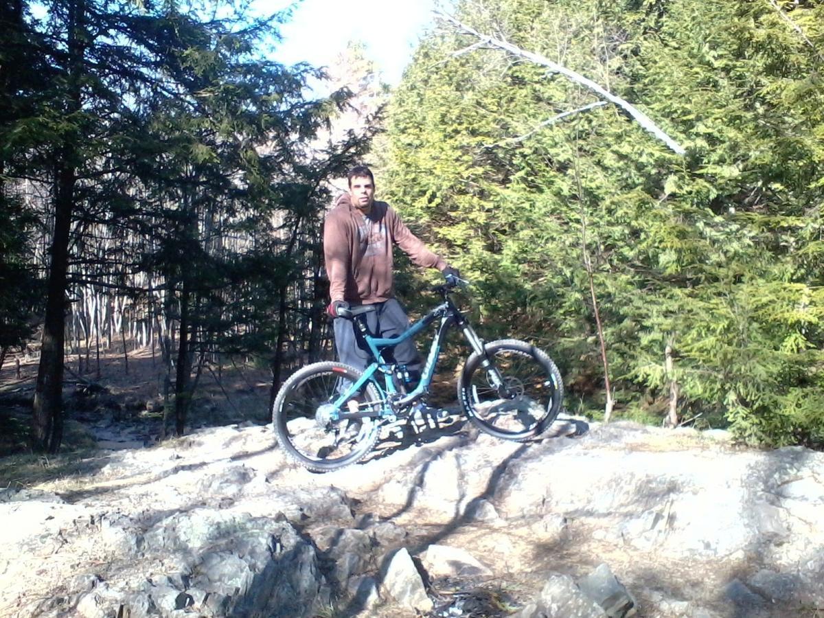 A person standing next to a mountain bike on rocky terrain, surrounded by green trees and a clear blue sky. The individual is wearing a brown hoodie and gray pants, posing confidently with the bike. Clifford Park mountain bike trail.