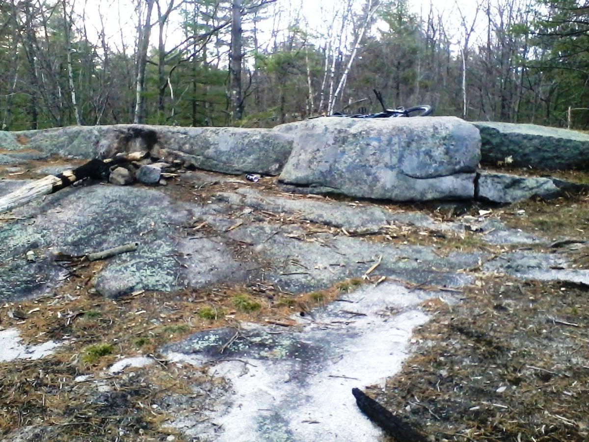 A rocky outdoor area featuring large boulders and a sandy patch. On the ground, there are remnants of a fire ring made of stones and logs. The surrounding landscape includes sparse trees and foliage, indicating a wilderness setting. Clifford Park mountain bike trail.