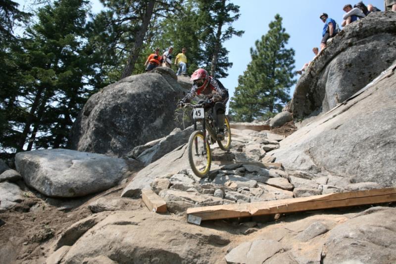 A mountain biker navigates a rocky downhill trail, wearing a helmet and protective gear. The rider is on a steep path made of large stones, with a wooden plank providing support. Spectators watch from above, surrounded by tall trees under a clear blue sky.