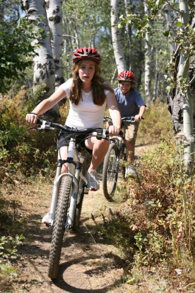 Two children riding mountain bikes along a narrow dirt trail surrounded by greenery and birch trees. The girl in the foreground, wearing a white tank top and a red helmet, has an expression of excitement as she rides. The boy in the background, wearing a blue shirt and a red helmet, is following her on the trail. The scene conveys a sense of adventure and outdoor activity.