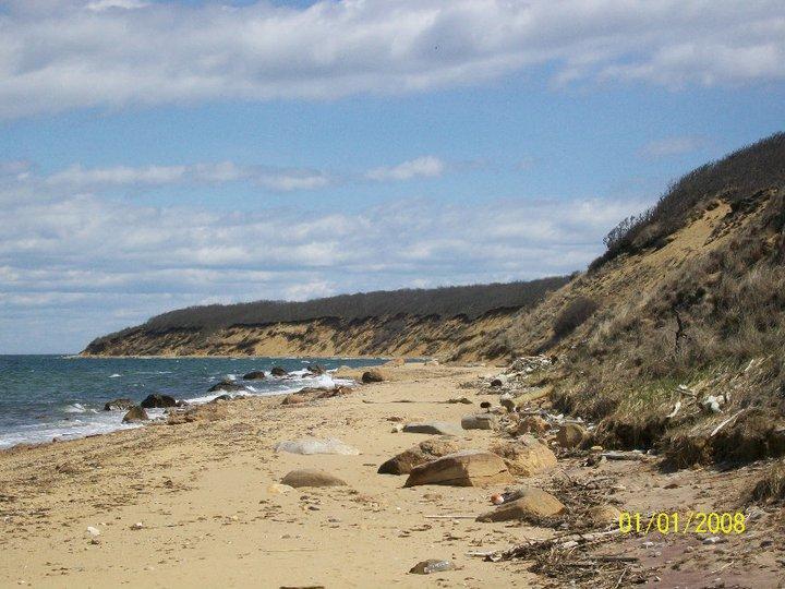 A scenic view of a sandy beach with a gentle slope leading to the ocean. The shoreline is dotted with rocks and debris, and a cliff rises in the background, partially covered by grassy vegetation. The sky is partly cloudy, adding a serene atmosphere to the coastal landscape. Hither Woods mountain bike trail.