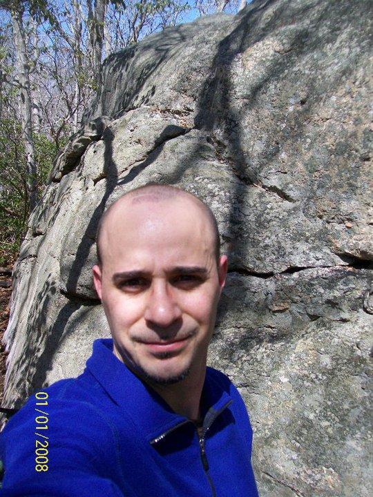 A person standing in front of a large rock formation, surrounded by trees. The individual is wearing a blue zip-up jacket and appears to be taking a selfie in a natural outdoor setting. Bright sunlight and shadows are visible, suggesting a clear day. Hither Woods mountain bike trail.