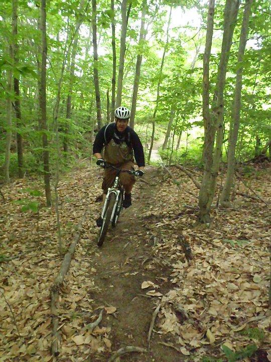 A mountain biker rides along a narrow trail in a lush forest, surrounded by green trees and scattered leaves on the ground. The cyclist wears a helmet and is focused on navigating the path. Bond Brook Trails mountain bike trail.