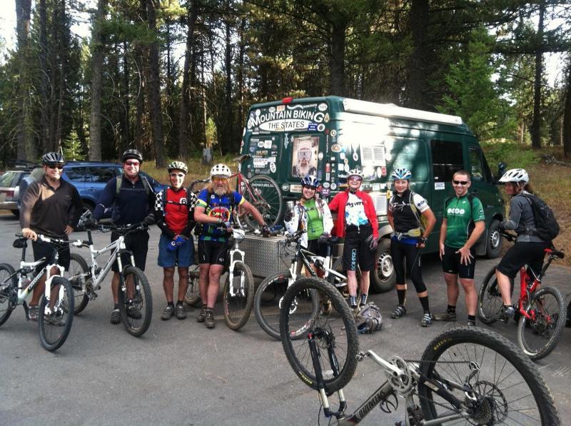 A group of mountain bikers poses for a photo near their parked van adorned with stickers and a mountain biking theme. The riders, wearing various cycling gear and helmets, are gathered in a forested area with trees in the background. Some bikes are leaning against the van, and the scene captures a sense of camaraderie and outdoor adventure.