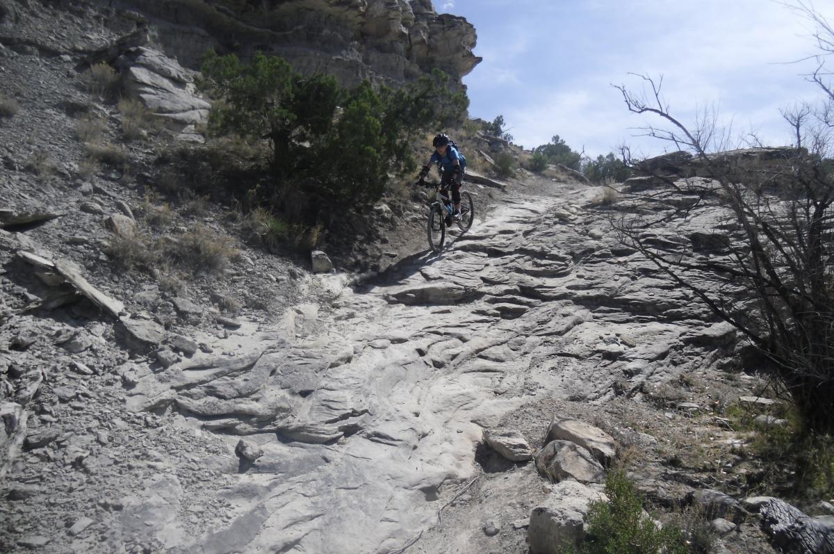 A mountain biker navigating a rocky trail on a hillside with sparse vegetation and gray rock formations under a clear sky. The Ribbon mountain bike trail.
