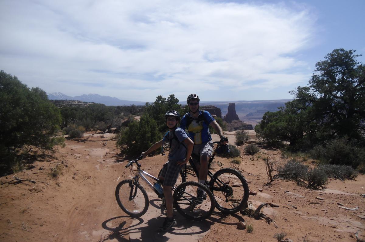 Two cyclists pose for a photo on a dirt trail amidst a scenic landscape, featuring green trees and distant mountains. The trail is sandy and surrounded by sparse vegetation, with rocky formations visible in the background. The cyclists are wearing helmets and casual athletic clothing. The sky is partly cloudy, suggesting a bright, sunny day. Dead Horse Point State Park mountain bike trail.