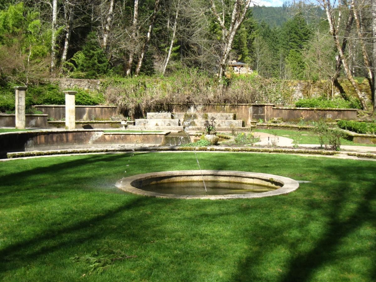 A serene garden featuring a circular stone fountain surrounded by lush green grass. In the background, stone walls and terraced landscaping are visible, with trees and shrubs providing a natural backdrop. The scene is illuminated by natural sunlight, giving a peaceful and tranquil atmosphere. Mckenzie River Trail mountain bike trail.