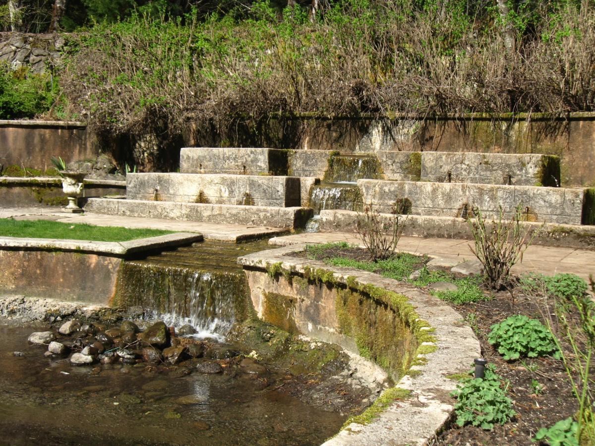A serene garden scene featuring a stone water feature with cascading waterfalls over steps, surrounded by lush greenery. The water flows into a small pond bordered by rocks, and decorative plants are visible throughout the area, enhancing the natural ambiance. Mckenzie River Trail mountain bike trail.