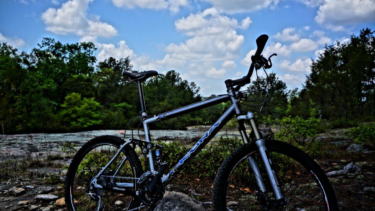 A silver mountain bike leaning against a rock in a natural setting, surrounded by green trees and a cloudy blue sky. The ground is rocky and uneven, with patches of grass and shrubs. Georgia International Horse Park mountain bike trail.
