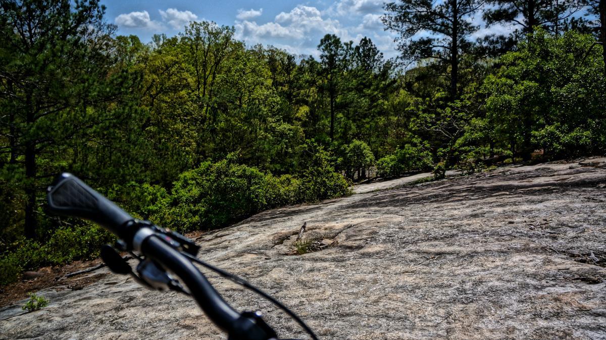 A mountain bike handlebar in the foreground, overlooking a rocky terrain and lush green trees under a partly cloudy blue sky. Georgia International Horse Park mountain bike trail.