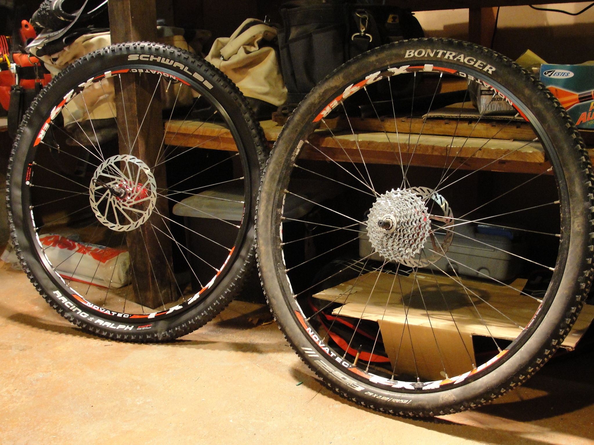 A pair of mountain bike wheels resting on a garage floor, featuring black rims with white and red accents. The wheels are equipped with disc brakes and have Schwalbe tires. In the background, various tools and bags are visible on shelves, suggesting a bike maintenance setting.
