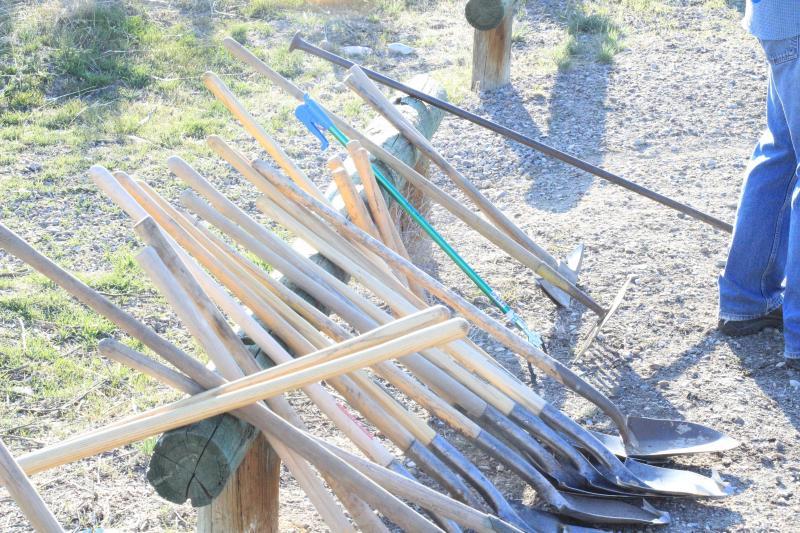 A collection of various garden tools, including shovels and hoes, leaning against a wooden post on a gravel path, with a person partially visible in blue jeans in the background. The scene is outdoors, set in a grassy area with sunlight illuminating the tools.