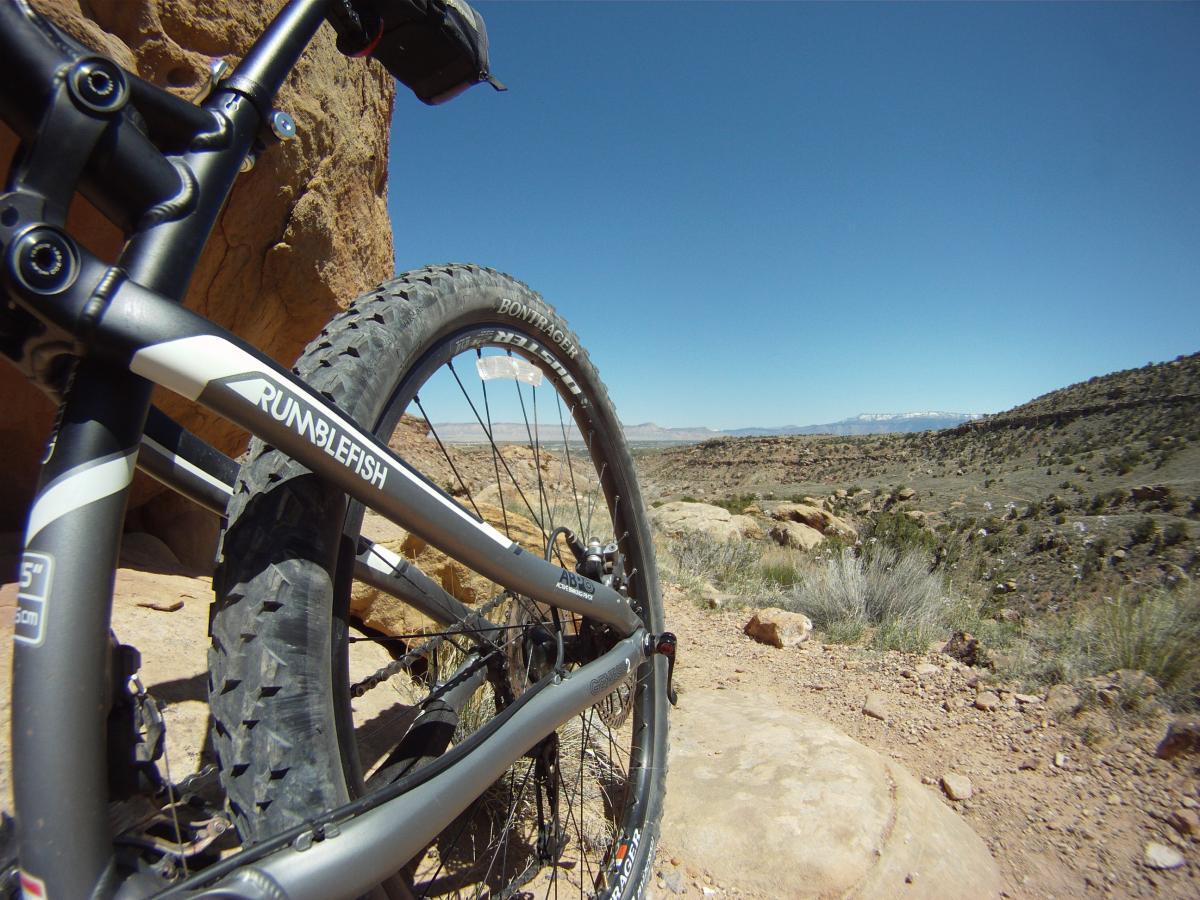 Close-up view of a mountain bike resting on rocky terrain, with the landscape stretching into the distance under a clear blue sky. The bike frame is partially visible, featuring the name "Rumblefish" along the side, and the tire shows tread patterns suitable for off-road riding. The surrounding environment includes rocky outcrops and sparse vegetation, typical of a rugged, outdoor setting. Gunny Loop mountain bike trail.