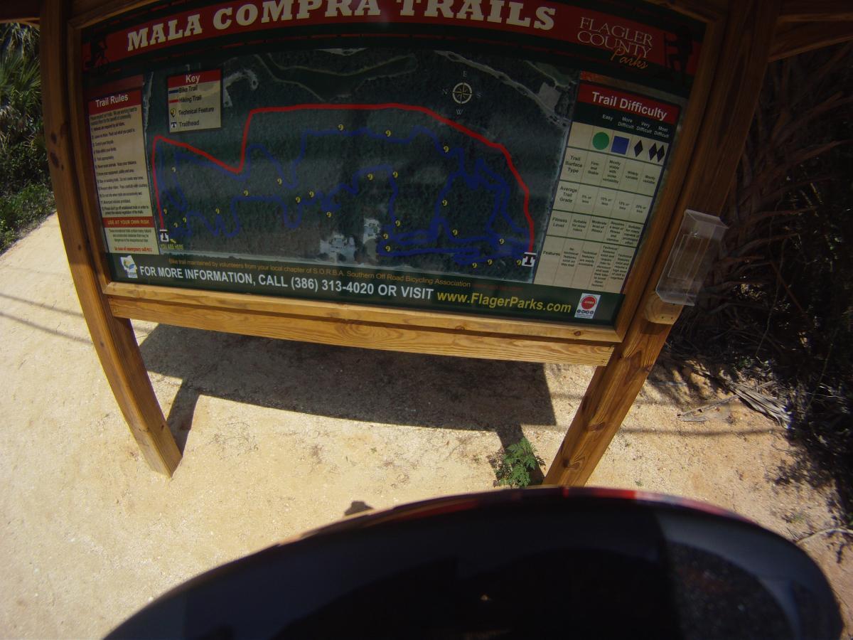 A wooden sign displaying a map of the Mala Compra Trails, located in Flagler County. The map includes trails for biking and walking, marked with different colors to indicate difficulty levels, along with key features and a compass. The ground is sandy, and part of a helmet is visible in the foreground. Mala Compra mountain bike trail.