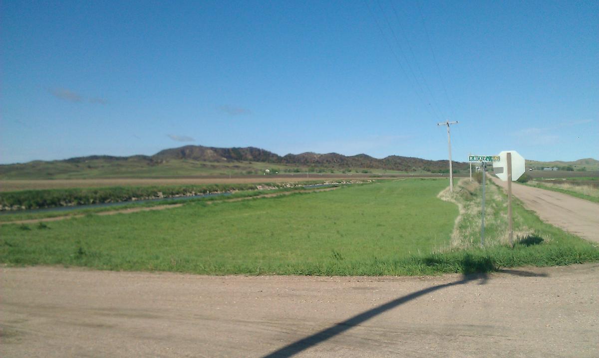 A scenic view of a rural landscape featuring a wide green field, a winding creek, and distant mountains under a clear blue sky. A dirt road intersects the foreground, with a road sign visible on the right side indicating a nearby location. Power lines stretch along the side of the road. Potters Pastures mountain bike trail.