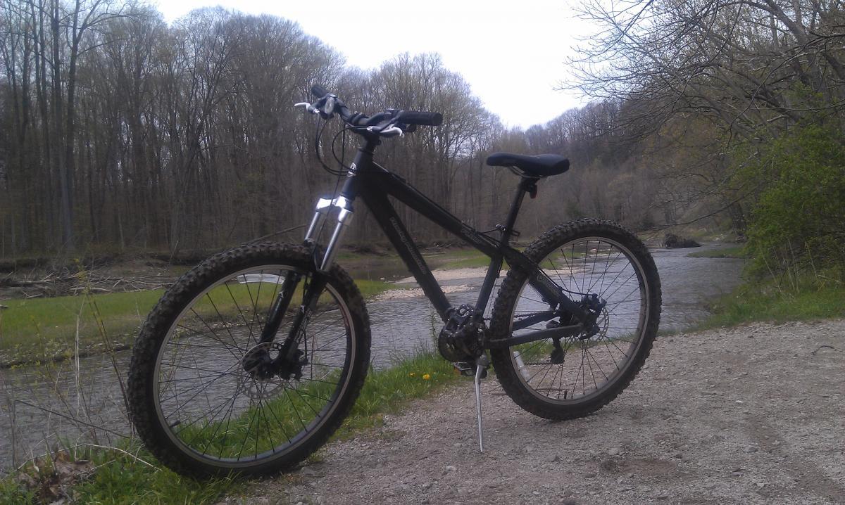 A black mountain bike parked on a gravel path alongside a river, surrounded by trees and greenery in a natural setting. Atchinson (hogback Ridge North) mountain bike trail.