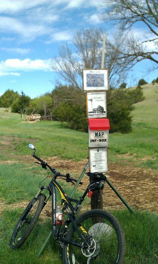 Image of a mountain bike leaning against a wooden post with an information box. The box has a map and various notices. In the background, there are green fields and trees under a partly cloudy sky. The scene suggests a recreational area for biking or hiking. Potters Pastures mountain bike trail.