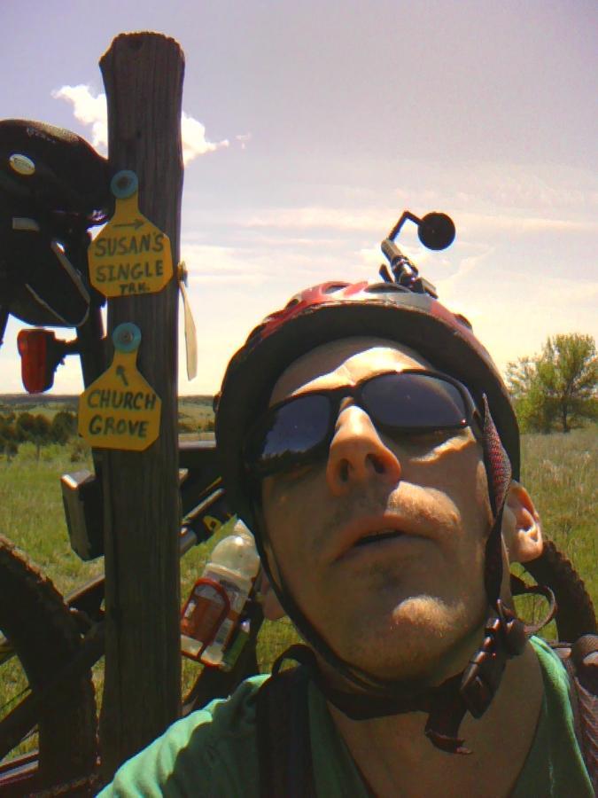 A person wearing sunglasses and a helmet takes a selfie in front of a post with trail signs labeled "Susan's Singletrack" and "Church Grove." A mountain bike is partially visible behind them, and the landscape features open grassland under a clear blue sky. Potters Pastures mountain bike trail.