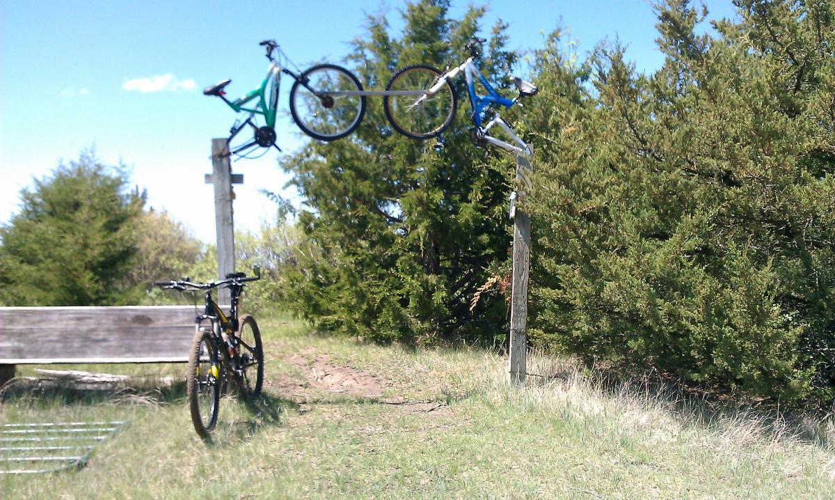 Two bicycles are suspended from a wooden post, with one bike in green and the other in blue. In the foreground, there is a black bicycle resting on the ground beside a wooden bench. The scene is set in a grassy area surrounded by trees under a clear blue sky, suggesting an outdoor park or trail environment. Potters Pastures mountain bike trail.