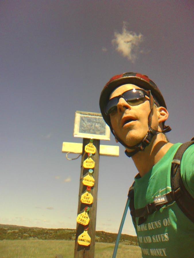 A person wearing a helmet and sunglasses stands near a trail marker post with colorful signs indicating various trail classifications. The sky is clear with a few clouds, and the landscape around them appears open and sunny. The individual is taking a selfie, looking slightly upwards with an expression of excitement or surprise. Potters Pastures mountain bike trail.