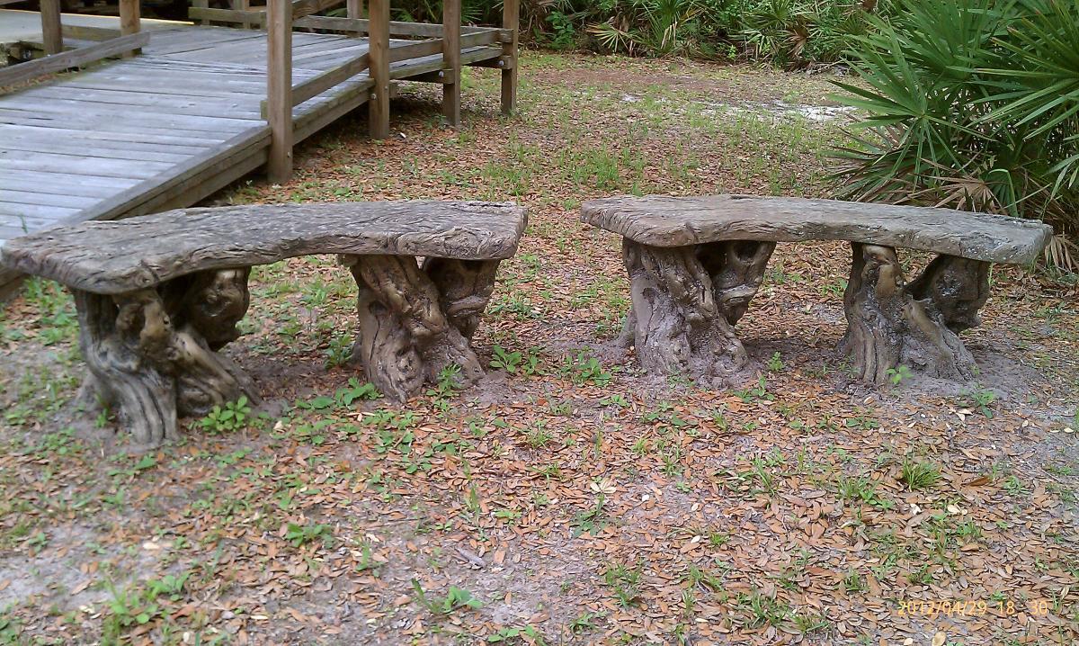 Two wooden benches with a natural, rustic design, placed on a grassy area surrounded by fallen leaves. The benches have curved tops and sturdy bases that resemble tree roots. A wooden deck is visible in the background, surrounded by lush greenery. Environmental Center mountain bike trail.