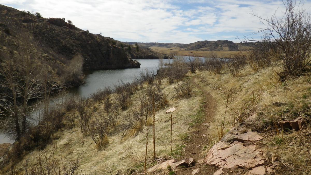 A scenic view of a winding dirt path along a riverbank, surrounded by sparse bushes and grassy terrain. In the background, there are hills and a cloudy sky, creating a tranquil natural landscape. Curt Gowdy State Park mountain bike trail.