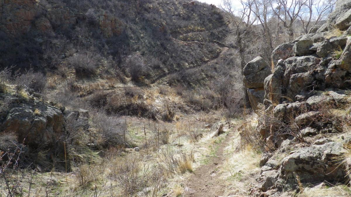 A rocky path winding through a dry, hilly landscape, surrounded by sparse vegetation and rocky formations under a clear sky. Curt Gowdy State Park mountain bike trail.