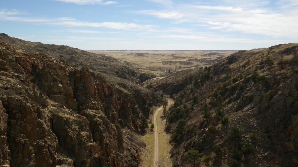 A vast landscape featuring rugged rocky cliffs and a winding dirt road below, set against a backdrop of rolling hills and a clear blue sky with scattered clouds. Curt Gowdy State Park mountain bike trail.