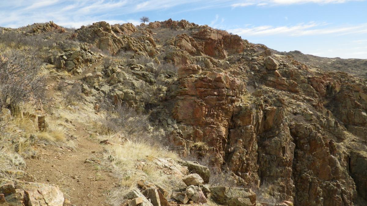 A rocky trail winding through rugged terrain, surrounded by sparse vegetation and steep rock formations under a partly cloudy blue sky. Curt Gowdy State Park mountain bike trail.