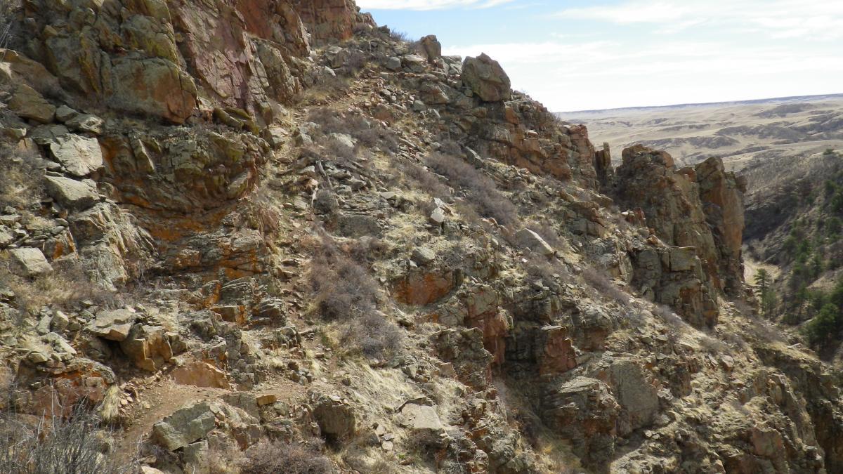 A rocky hillside with rugged terrain, featuring scattered boulders and sparse vegetation. The landscape includes a winding path leading up the slope, with distant rolling hills and a cloudy sky in the background. Curt Gowdy State Park mountain bike trail.