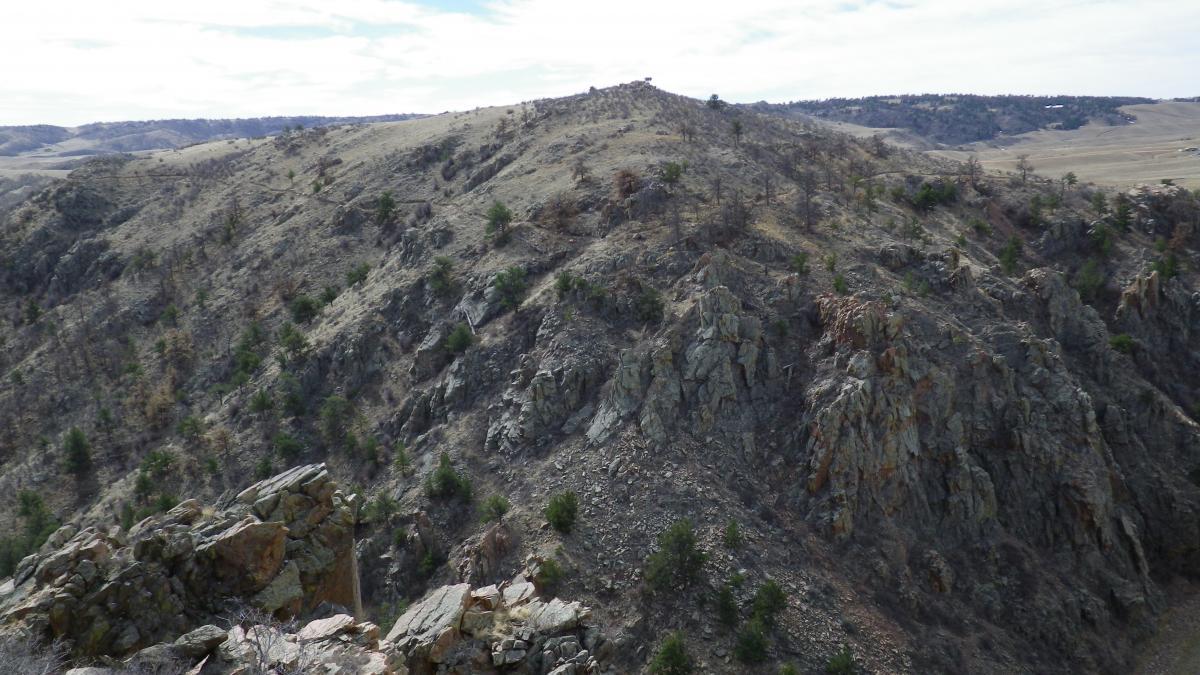 A scenic view of rugged hills and rocky terrain, featuring patches of green vegetation and sparse trees, under a partly cloudy sky. The landscape displays a mix of rocky outcrops and rolling hills, typical of a natural wilderness area. Curt Gowdy State Park mountain bike trail.