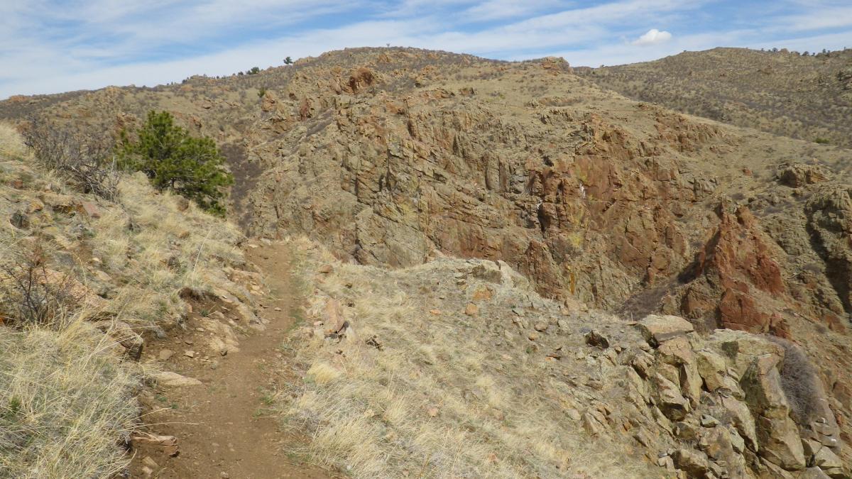 A narrow dirt path winds along a rocky ridge in a mountainous landscape. The terrain is dry and rugged, with patches of grass and sparse vegetation. In the distance, more rocky hills and a blue sky with scattered clouds can be seen, creating a serene outdoor setting. Curt Gowdy State Park mountain bike trail.