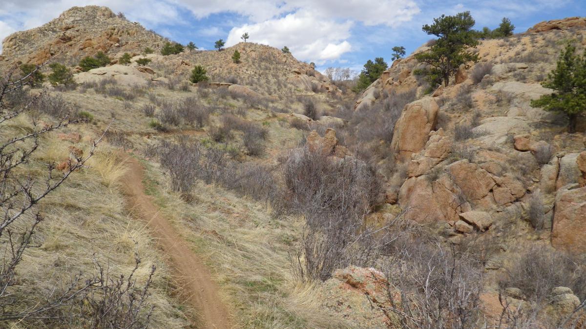 A winding dirt path leads through a rocky, semi-arid landscape with sparse vegetation. Rolling hills are dotted with small trees and shrubs, and the sky is partly cloudy, creating a contrasting backdrop to the earth-toned rocks and soil. Curt Gowdy State Park mountain bike trail.