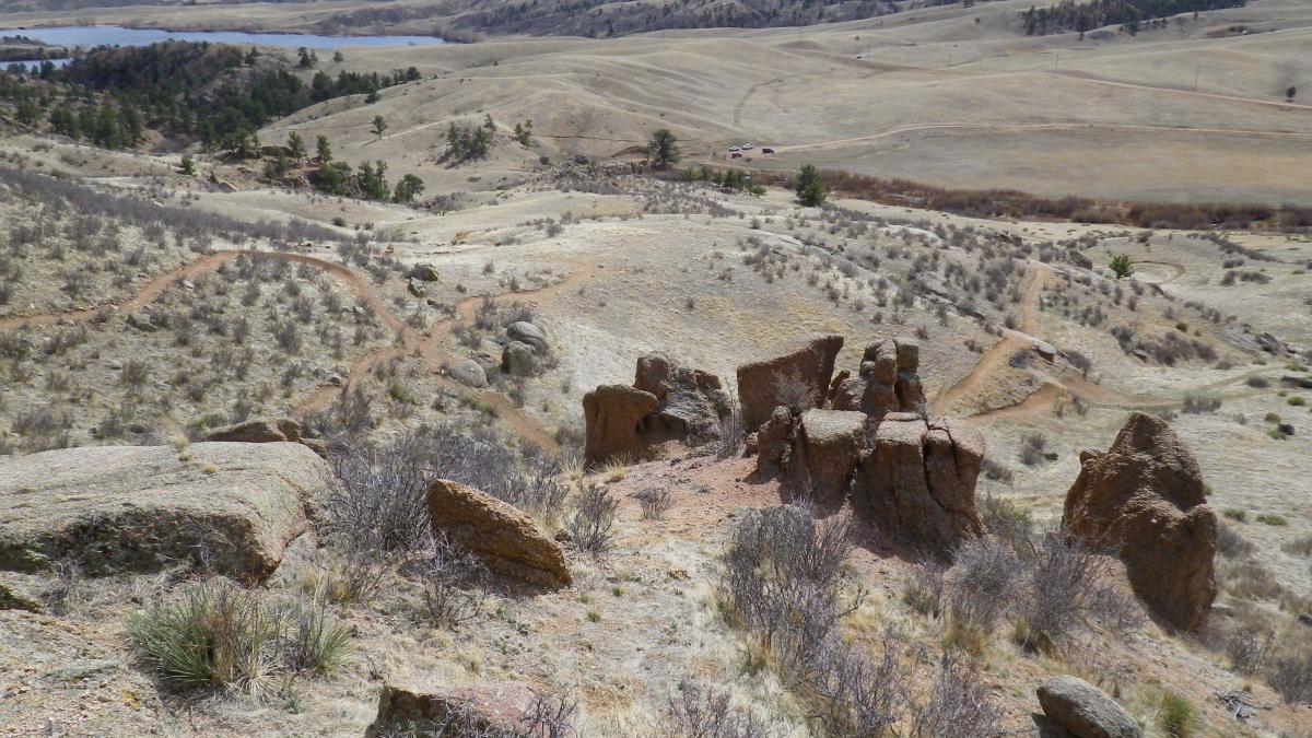 A scenic view of a hilly landscape featuring rocky outcrops, sparse vegetation, and winding trails. In the background, a calm body of water can be seen, surrounded by rolling hills and patches of trees. The area appears to be a natural outdoor setting, ideal for hiking and exploring. Curt Gowdy State Park mountain bike trail.