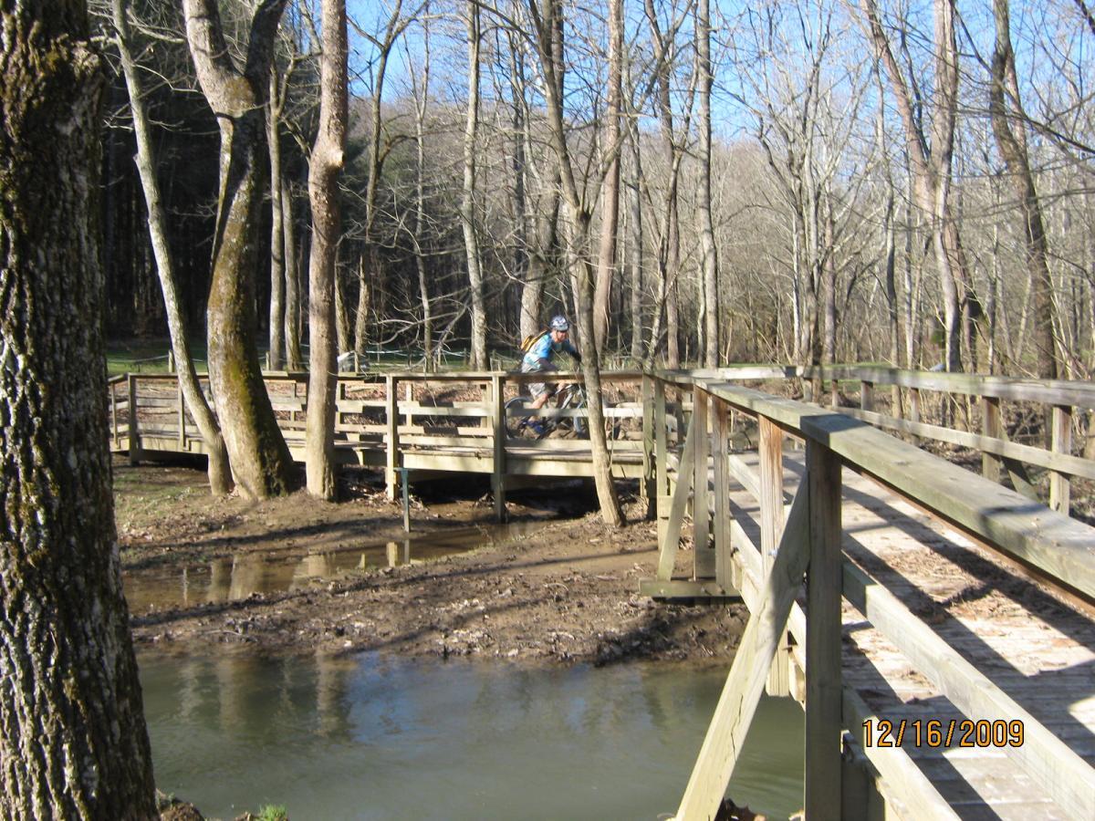 A cyclist riding over a wooden bridge in a wooded area, surrounded by trees that have lost their leaves. The bridge spans a small stream with muddy banks, and the scene is illuminated by bright sunlight on a clear day. Sugar Hollow Park mountain bike trail.
