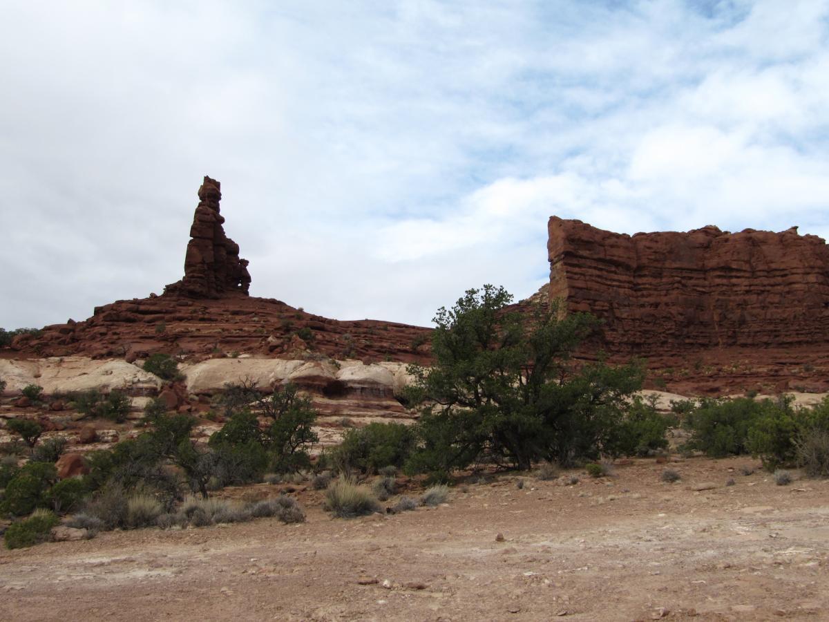 A rocky landscape featuring a prominent stone formation resembling a spire, with layers of reddish rock in the background. Sparse vegetation, including small bushes and shrubs, is visible on the sandy terrain under a cloudy sky. Teapot Rock - Standing Rock, Canyonlands national park mountain bike trail.