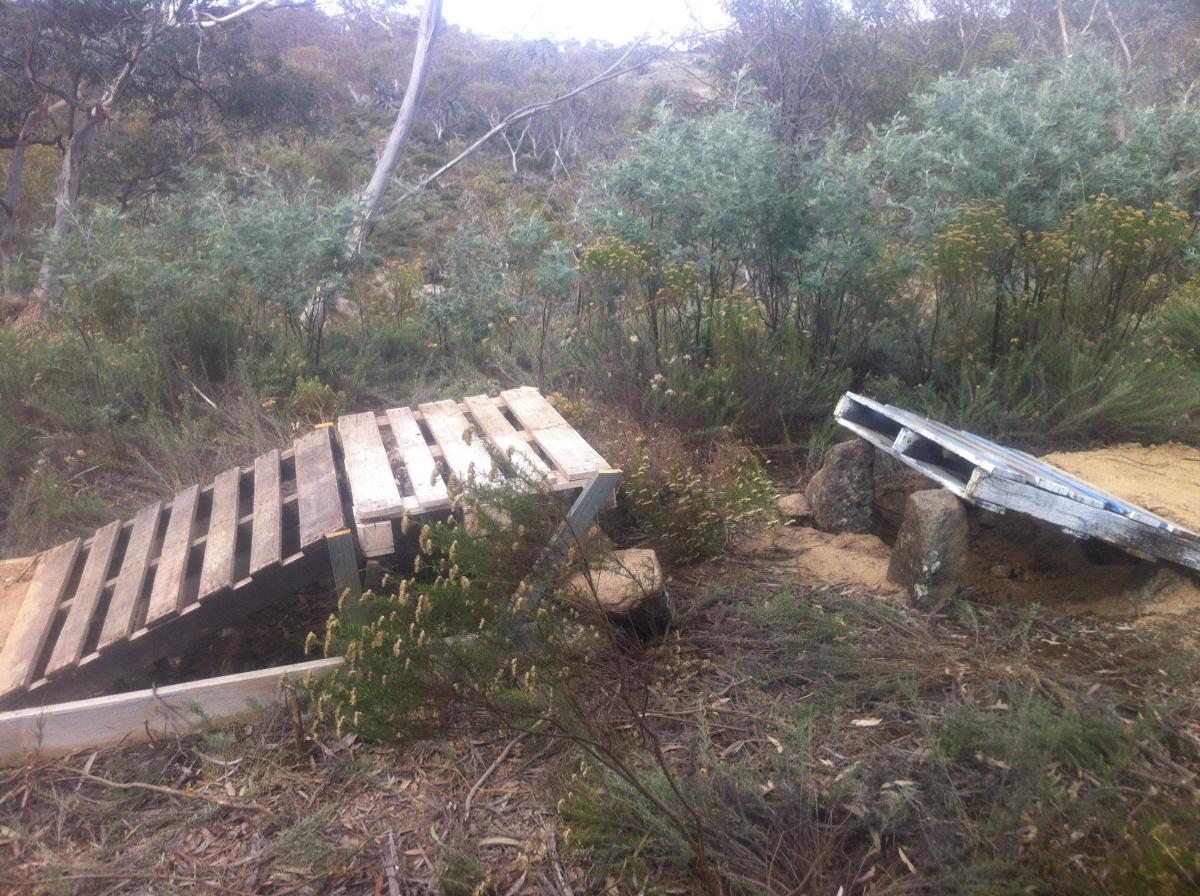 Two wooden pallets are positioned at an angle against rocky terrain, surrounded by low green vegetation and scattered rocks. The area appears natural and slightly rugged, typical of a bushland environment. East Jindabyne mountain bike trail.