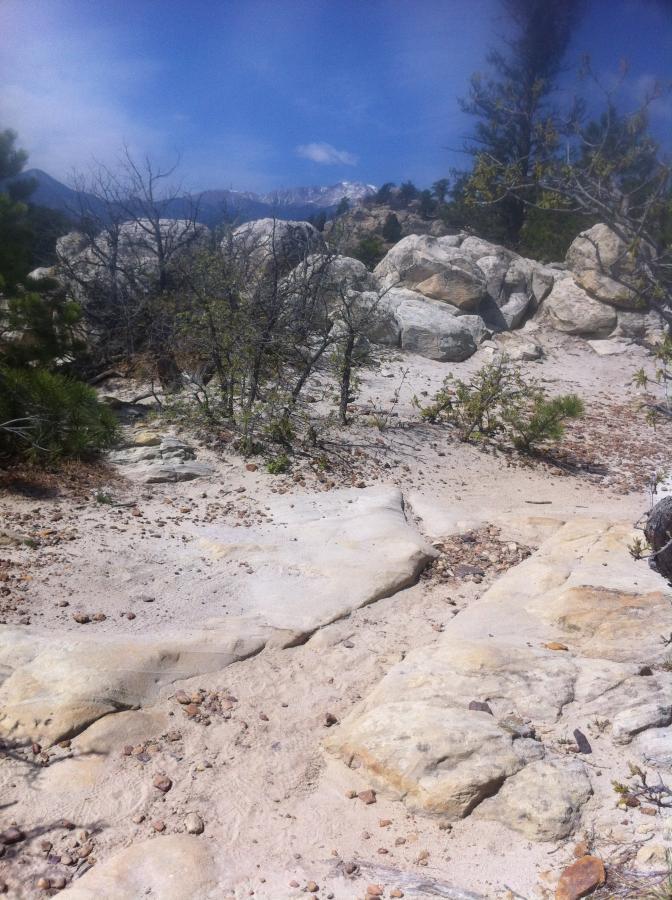 A rocky trail leading through a natural landscape, with scattered boulders, sparse vegetation, and mountains in the distance under a partly cloudy sky. Ute Valley Park mountain bike trail.
