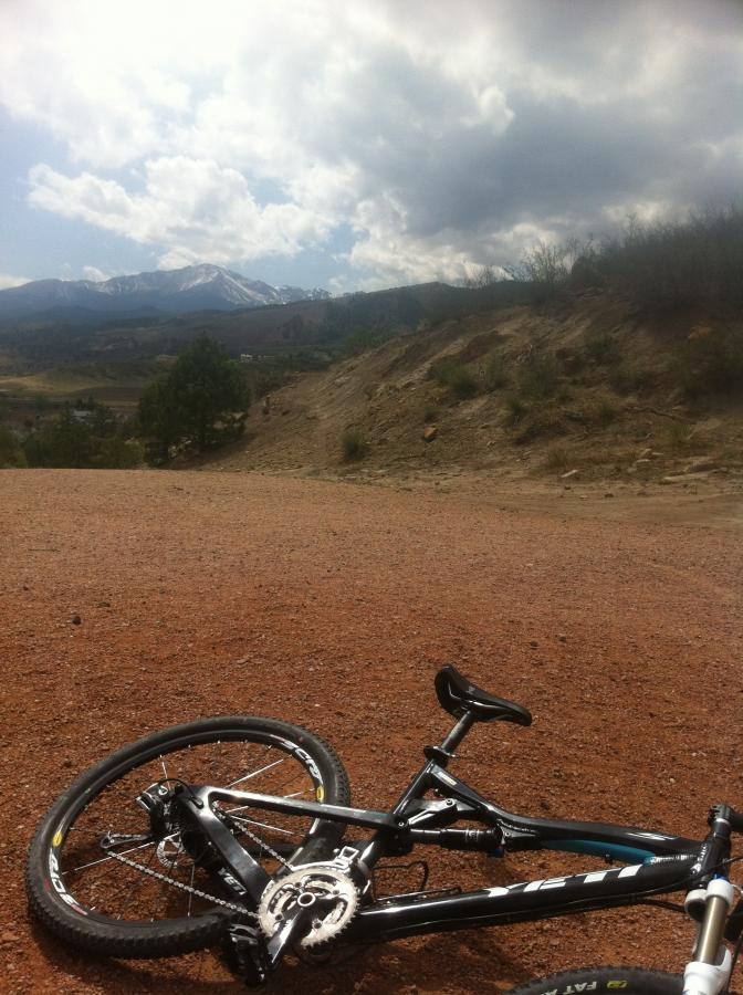 A mountain bike resting on gravel ground with a backdrop of cloudy skies and snow-capped mountains in the distance. Ute Valley Park mountain bike trail.