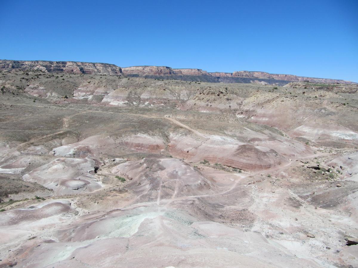 A panoramic view of a rugged desert landscape featuring multicolored rocky terrain with shades of pink, beige, and gray. The vast, dry ground leads to rocky cliffs in the background under a clear blue sky, showcasing the unique geological formations of the area. Gunny Loop mountain bike trail.