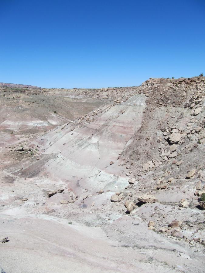 A wide view of a rugged landscape featuring layered rock formations in shades of beige, gray, and pink, under a clear blue sky. The ground is mostly dry and rocky, with scattered boulders and gently sloping hills. Gunny Loop mountain bike trail.