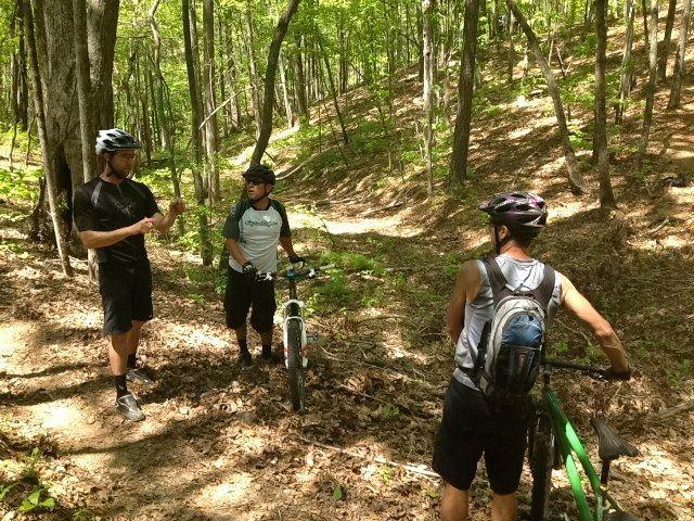 Three mountain bikers pause on a wooded trail, discussing their route. The scene is surrounded by tall trees with vibrant green leaves, and the ground is covered in fallen leaves. Two riders are standing near their bikes, while one is leaning on a bike, listening attentively. The atmosphere suggests a shared moment of camaraderie in nature. Woodring Branch mountain bike trail.