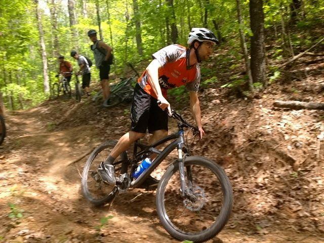 A mountain biker in a bright orange and gray jersey navigates a dirt trail surrounded by lush green trees, leaning into a turn. In the background, several other cyclists pause on their bikes along the trail. The scene captures an active outdoor moment in a forested area. Ridgeway Loop mountain bike trail.