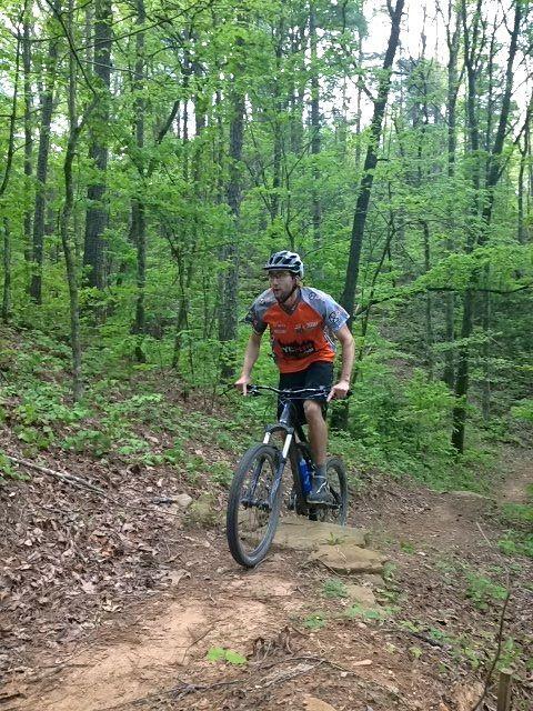 A mountain biker riding on a dirt trail through a lush green forest. The rider is wearing a helmet and an orange and gray jersey, focused on navigating the terrain. Surrounding trees and foliage indicate a vibrant outdoor setting. Ridgeway Loop mountain bike trail.