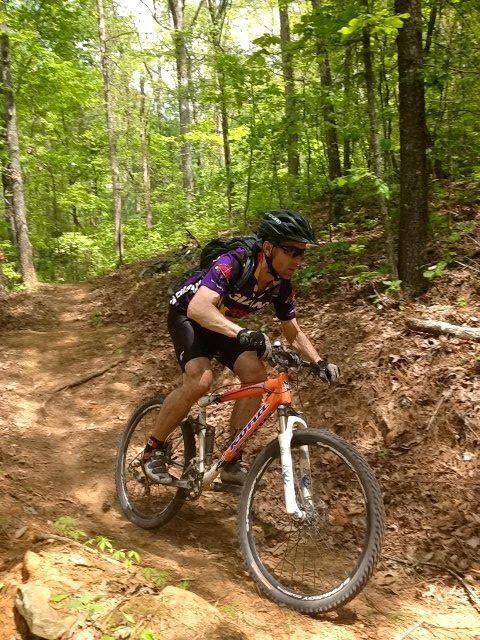 A mountain biker navigates a dirt trail in a lush, green forest. The rider is wearing a helmet and a colorful cycling jersey, leaning forward as they pedal along the winding path surrounded by trees and foliage. Ridgeway Loop mountain bike trail.