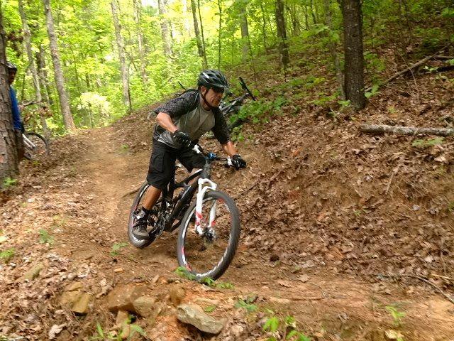 A person riding a mountain bike on a winding dirt trail surrounded by trees. The cyclist is wearing a helmet and protective gear, leaning into the turn while navigating the terrain. The scene is set in a lush, green forest with fallen leaves on the ground. Ridgeway Loop mountain bike trail.