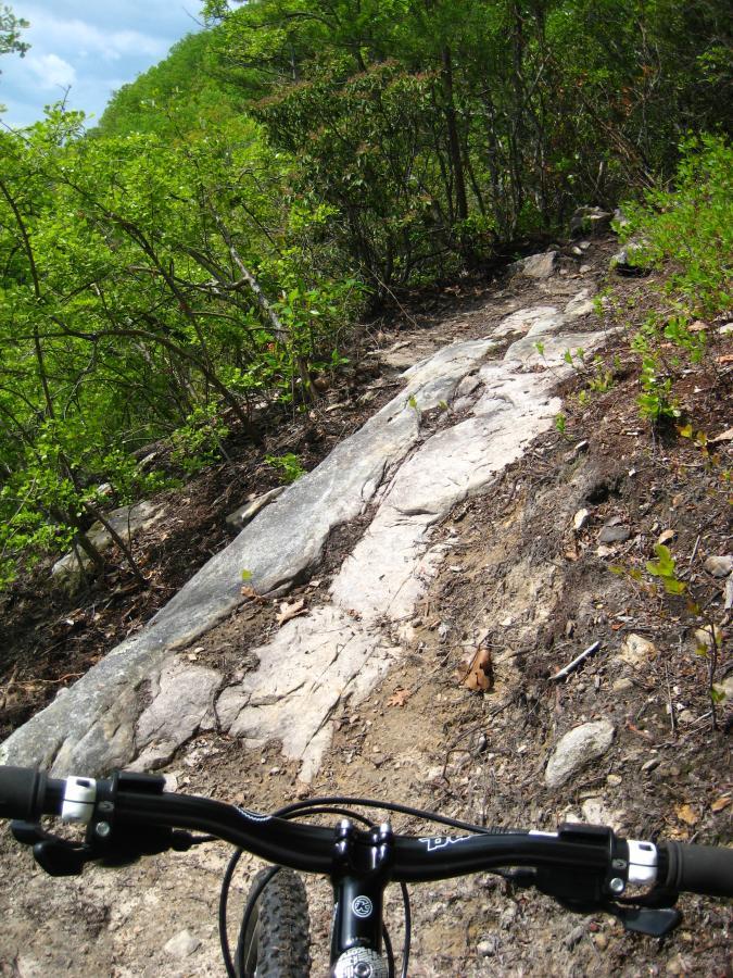 A mountain bike handlebar is visible in the foreground, pointing down a rocky trail surrounded by lush greenery. The path is uneven, featuring exposed rocks and dirt, indicating a challenging terrain for biking. The sky above is partly cloudy, adding to the outdoor adventure atmosphere. Windrock Bike Park mountain bike trail.